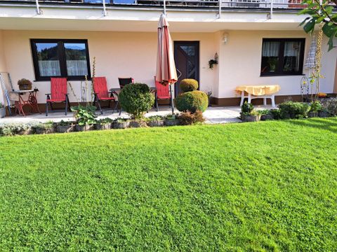 Een verzorgde tuin met groen gras en verschillende planten. Voor het huis staan ​​tuinstoelen met kussens en een parasol.