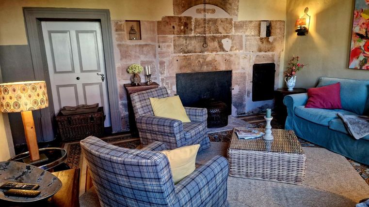 A cozy living room with a fireplace, two armchairs, and a couch. The walls are painted in warm colors and there are decorative elements such as flowers and a rug.