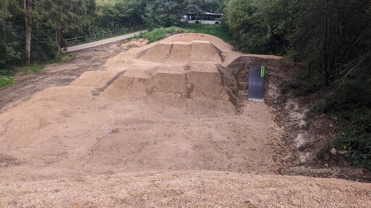 Eine sandige Sprungschanze in einem Waldgebiet. Im Hintergrund sind einige Bäume und ein Weg sichtbar.