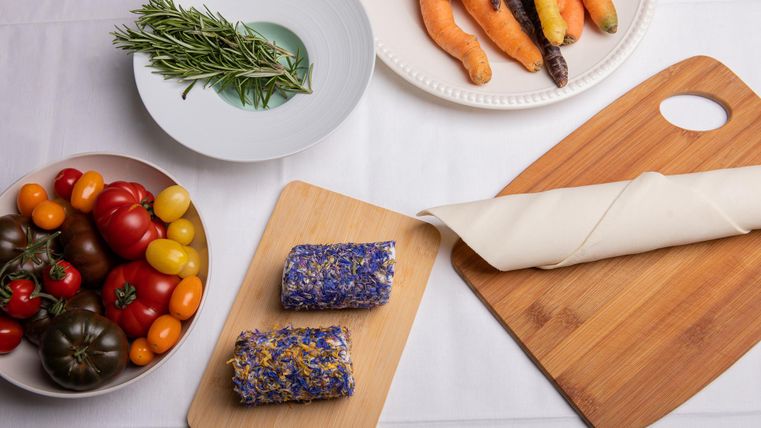 Een kleurrijke selectie van verse groenten en tomaten is te zien. Ook zijn er twee decoratieve stukken geitenkaas op een houten plank te herkennen.