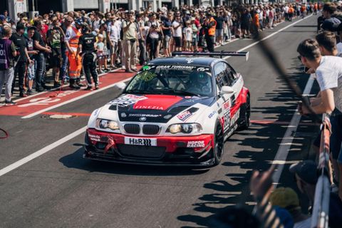 Ein Rennwagen fährt auf einer Rennstrecke vorbei, umgeben von zahlreichen Zuschauern. Die Atmosphäre ist lebhaft und voller Spannung.