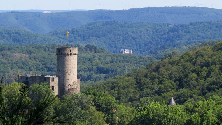 Blick über bewaldete Hügel mit Burg Pyrmont im Vordergrund, erkennbar am runden Turm mit Fahne. In der Ferne ist Burg Eltz zwischen den Waldhängen zu sehen.