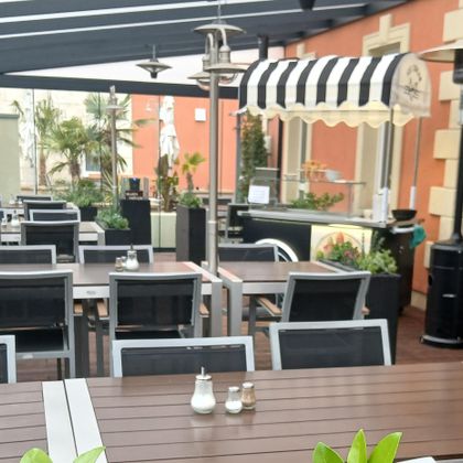 Ein gemütlicher Außenbereich eines Restaurants mit modernen Tischen und Stühlen. Es gibt Pflanzen und Sonnenschirme, die eine einladende Atmosphäre schaffen.