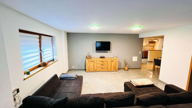Een moderne woonkamer met een zwarte bank en een houten kast. Aan de muur hangt een televisie en er zijn grote ramen met jaloezieën.