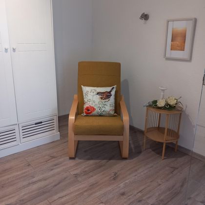 Een comfortabele stoel met een kleurrijk kussen staat in een lichte kamer. Naast staat een kleine tafel met bloemen en een decoratie.