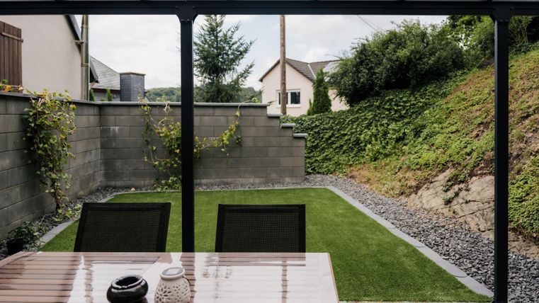 Un espace extérieur couvert et confortable avec une table et des chaises de jardin. En arrière-plan, on peut voir le petit jardin récemment aménagé.
