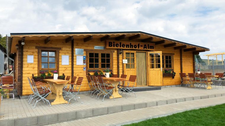 <p>In the background, you can see a large, single-story wooden hut. In the middle, there is a double door. Above it hangs a wooden sign with the inscription "Bielenhof Alm". On the right and left of the door, there are casement windows with flower boxes. In front of the hut, there is a paved terrace with some tables and chairs. In the foreground, there is a grassy area.</p>