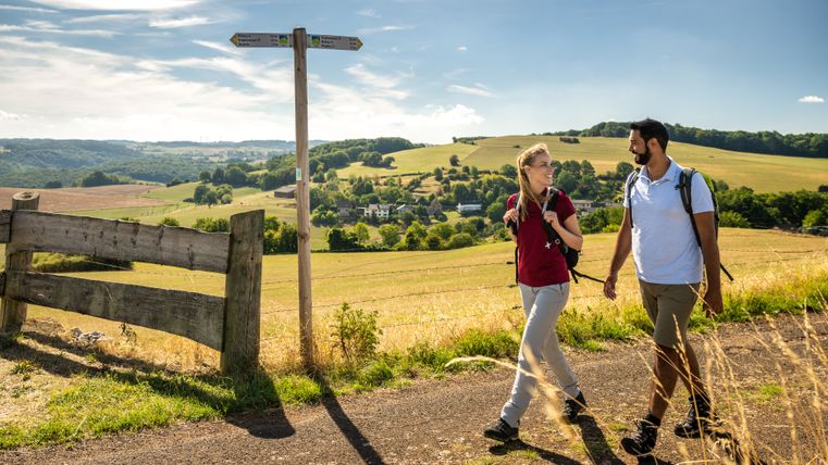 Two hikers on a path in the Eifel with a signpost and a wide view over the landscape.