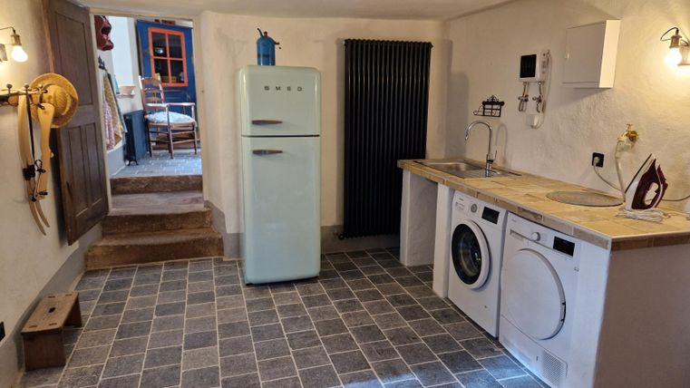A modern kitchen with gray tiles, a refrigerator, and a washing machine. In the background, furniture and a door to another room are visible.