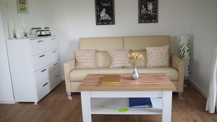 Ein helles Wohnzimmer mit einem beigen Sofa und einem Holztisch. An der Wand hängen zwei Bilder, und der Raum ist mit einer modernen Kommode und Pflanzen dekoriert.