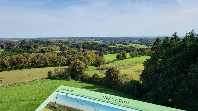 Panoramic view from the Weißer Stein viewing tower over green landscapes and forests with information board in the foreground.