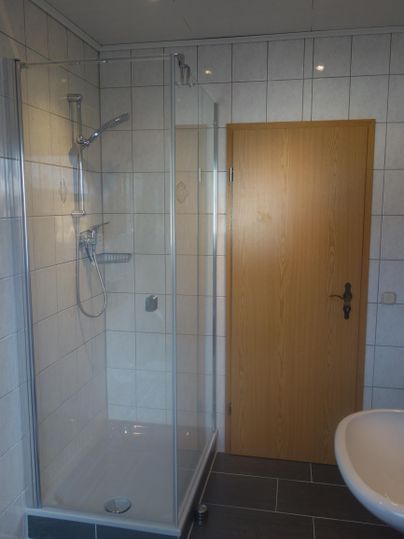 A modern bathroom with a glass shower cabin and a sink. The walls are covered with white tiles and a wooden interior door is visible.