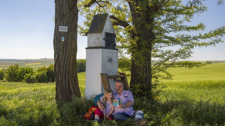 A man and a child sit under a wayside shrine on Paradiesweg in Polch, surrounded by green fields and trees.