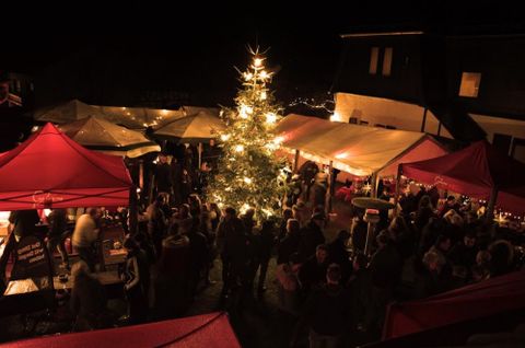 Un marché de Noël festif la nuit avec beaucoup de monde. Au centre se trouve un arbre de Noël illuminé et des auvents rouges.
