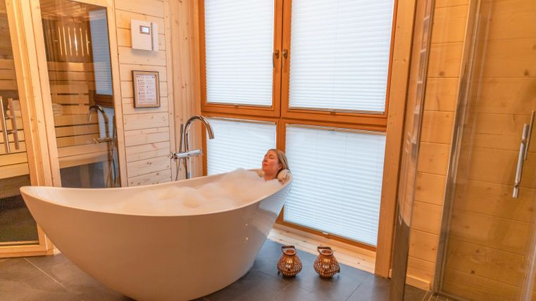 Eine entspannende Badewanne in einem modernen Badezimmer aus Holz. Eine Frau genießt ein Bad mit Schaum und Blick durch große Fenster.