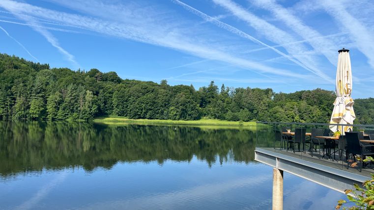 Stausee Bitburg mit Terrasse und Sonnenschirm, umgeben von Wald und blauem Himmel mit Kondensstreifen.