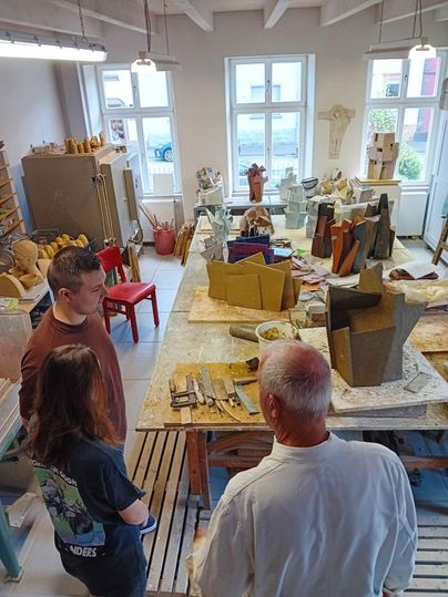 Ein Atelier mit mehreren Skulpturen und Werkzeugen. In der Mitte stehen zwei Personen im Gespräch.