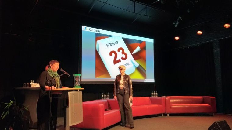 Een presentatie op een podium met een kalendervel dat 23 februari toont. Twee personen staan op het podium voor rode sofa's.