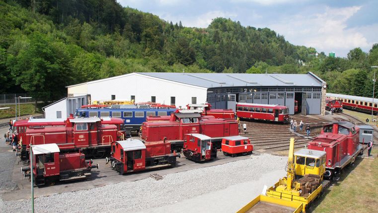 Eine Werkstatt für Züge mit vielen roten Lokomotiven und Bahnwaggons. Im Hintergrund sind grüne Bäume und ein blauer Himmel zu sehen.