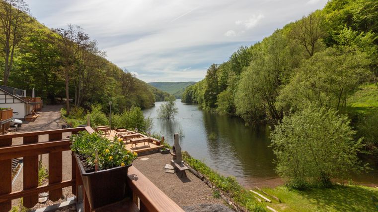 Une rivière calme, entourée de verdure luxuriante et d'arbres. Au premier plan, on aperçoit des terrasses en bois et des jardinières.