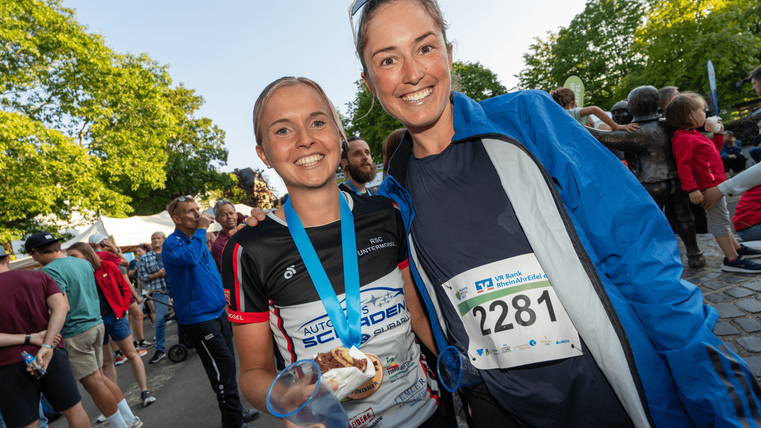 Zwei glücklich Auszeichnungen erhaltene Frauen stehen zusammen auf einem Sportevent. Im Hintergrund sind viele Menschen und Bäume zu sehen.