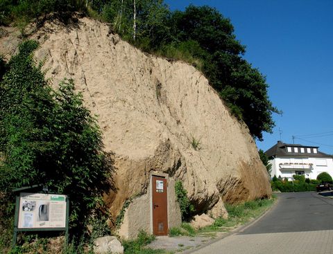 Ein steiler Hang mit sandiger Erde und Vegetation. Auf der linken Seite steht eine Tür, und im Hintergrund sind Gebäude zu sehen.
