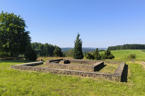 Ruïnes van een oud gebouw op een groene weide, omringd door bomen en heuvels onder een heldere, blauwe lucht.