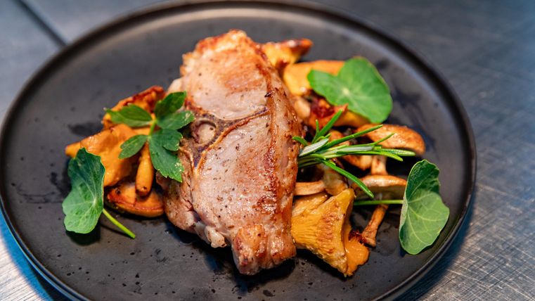 Ein gebratenes Stück Fleisch auf einem schwarzen Teller, umgeben von gebratenen Pilzen und frischen Kräutern. Das Gericht sieht appetitlich und sehr ansprechend aus.