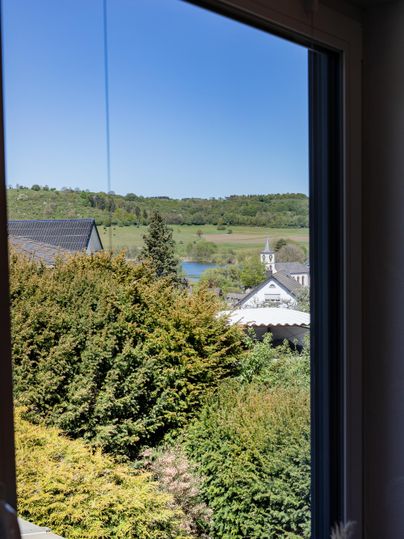 View from a window overlooking a green landscape with trees and a small lake. In the foreground, bushes and houses are visible.
