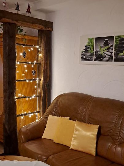 A cozy living room corner with a brown sofa and cushions. In the background, lights are hanging and a decoration with plant pictures is visible on the wall.