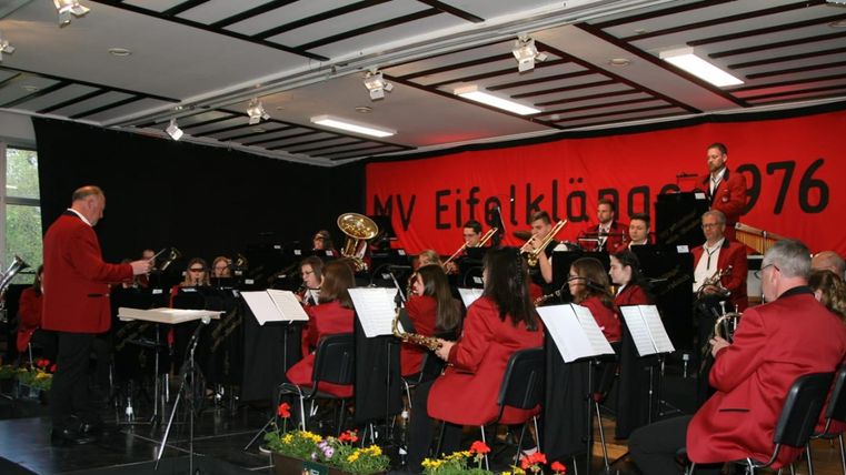 Eine Blaskapelle spielt auf einer Bühne. Die Musiker tragen rote Jacken und sind vor einem großen Banner mit dem Schriftzug "MV Eifelklingen 1976 e.V." aufgestellt.