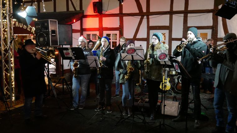 A band is playing music in a festively lit room. The musicians are standing together and playing various wind instruments.