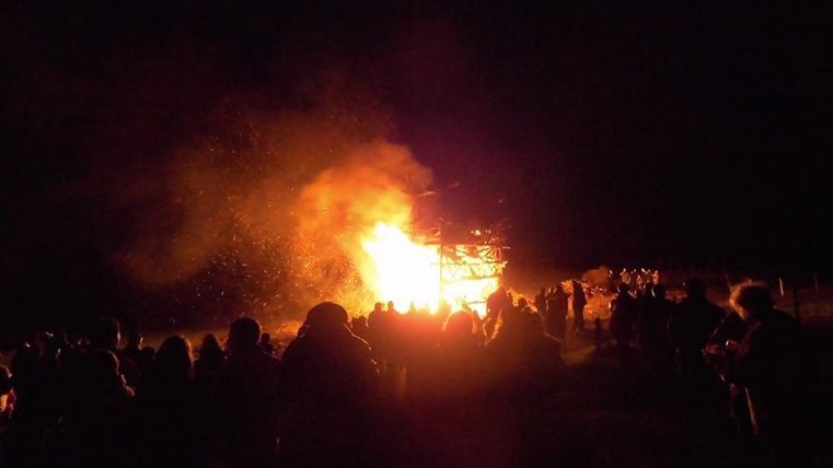 Ein großes Feuer brennt in der Nacht, umgeben von einer Menschenmenge. Funken fliegen in die Luft und schaffen eine festliche Atmosphäre.