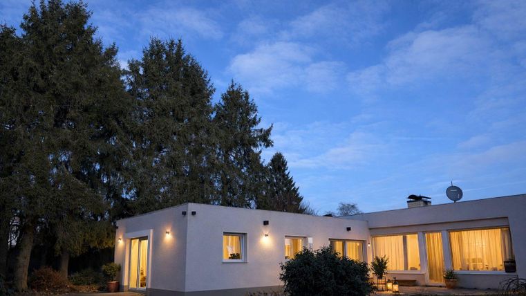 Ein modernes Haus umgeben von hohen Bäumen bei Dämmerungslicht. Die Fenster sind hell erleuchtet und schaffen eine einladende Atmosphäre.