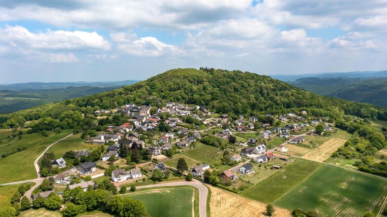 Een pittoresk landschap met een klein dorp aan de voet van een bosrijke heuvel. Op de achtergrond zijn groene velden en zachte heuvels zichtbaar.