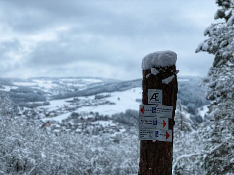 Verschneiter Wanderweg mit Wegweiser im Vordergrund und winterlicher Landschaft im Hintergrund.