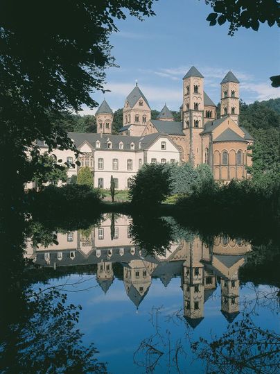 Een indrukwekkend gebouw met torens weerspiegelt zich in het rustige water van een meer. Omgeven door bomen en een schilderachtig landschap.