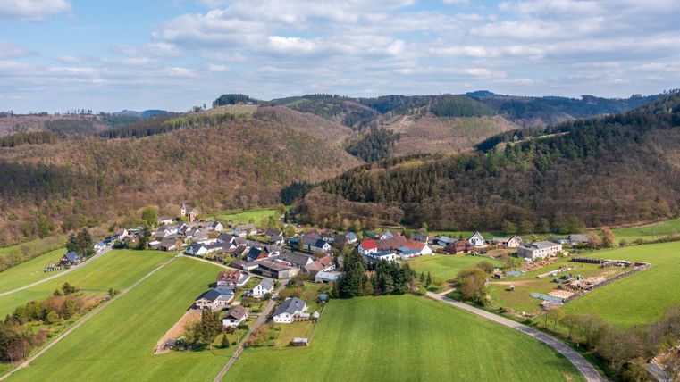 Eine malerische Dorfansicht umgeben von sanften Hügeln und grünen Feldern. Der Himmel ist klar mit einigen Wolken.