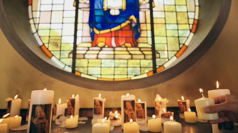 Burning candles with images of the Virgin Mary in front of a colorful church window with a religious motif; a hand adds another candle.
