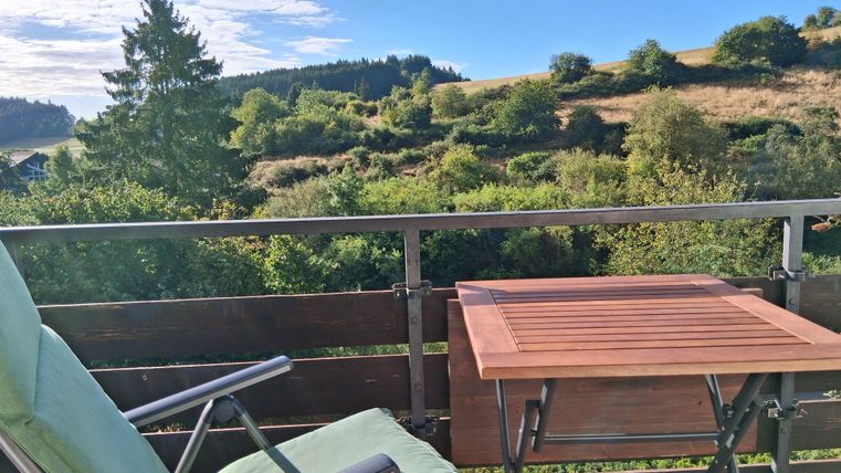 Een rustige balkon met een comfortabele stoel en een tafeltje. Op de achtergrond zijn groene heuvels en een blauwe lucht te zien.