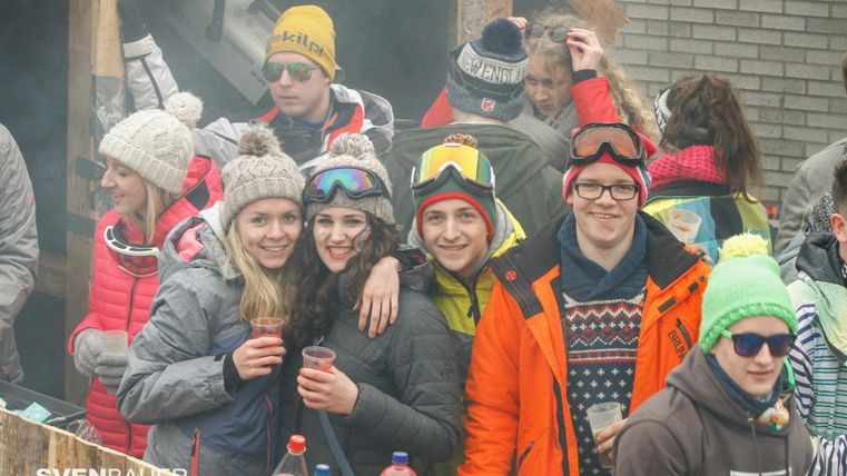 Eine Gruppe von jungen Menschen feiert im Freien auf einem Skigelände. Sie tragen warme Kleidung und Mützen, während sie Drinks in den Händen halten.