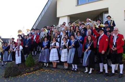 Eine große Gruppe von Menschen steht in traditionellen Trachten vor einem Gebäude. Viele halten Musikinstrumente, und die Stimmung ist festlich.