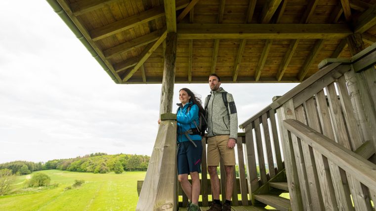 Twee mensen staan op een houten uitkijktoren en staren in de verte over een groen landschap.