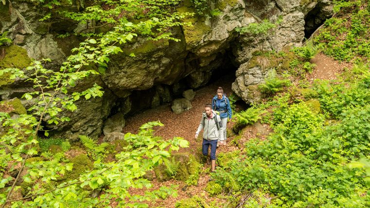 Two hikers walk past a rocky cave on a green forest path. Lush vegetation surrounds the trail.