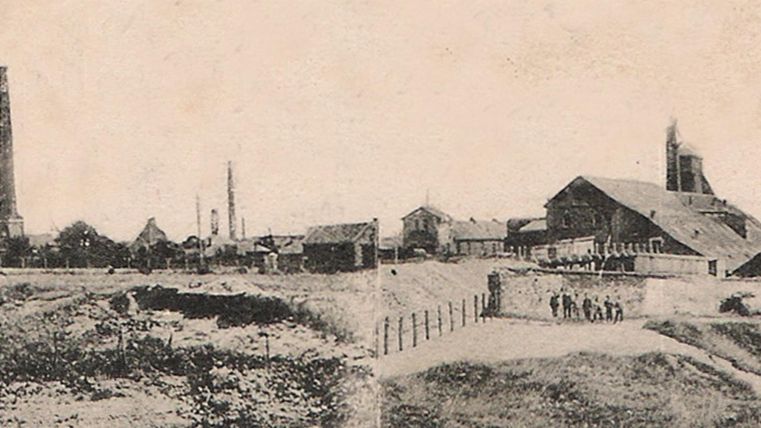 Une vue industrielle historique avec des bâtiments d'usine et de hauts cheminées. Au premier plan, on peut voir un paysage inégal avec des clôtures et quelques personnes.