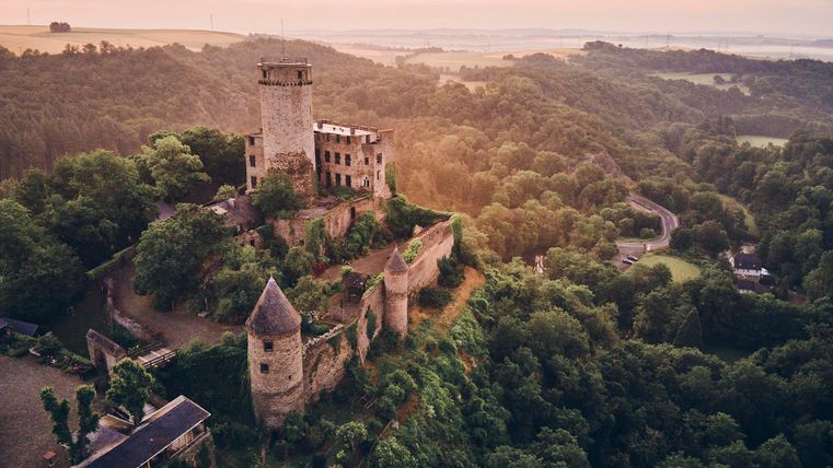 An old castle on a hill, surrounded by green forests. The morning sun illuminates the ruins and the landscape.