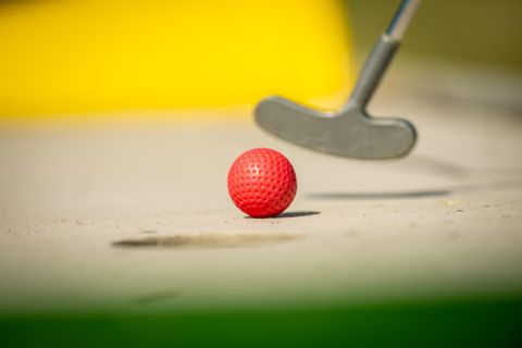 Ein roter Golfball liegt auf dem Boden. Ein Putter ist im Vordergrund sichtbar.