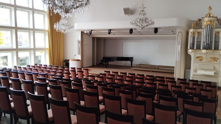 Ein großer Saal mit vielen Stühlen und einer Bühne. Es gibt Kronleuchter und ein Klavier auf der Bühne.