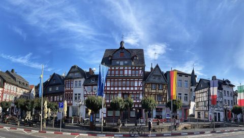 Een mooi historisch stadsplein met verschillende vakwerkhuizen. De lucht is helder en blauw, en er wapperen vlaggen aan de gebouwen.