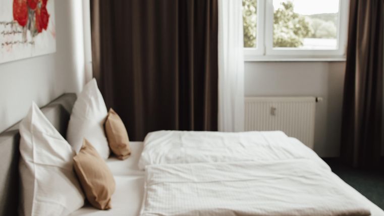 Une chambre confortable avec un lit double et des coussins doux. Les fenêtres sont équipées de rideaux et laissent entrer la lumière du jour.